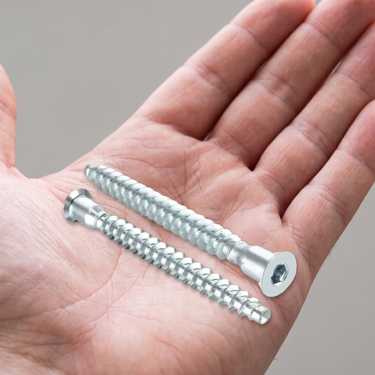 Two confirmat screws with a bright zinc-plated finish, shown in an open hand for scale